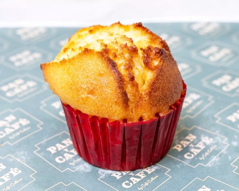 Golden-brown muffin in a red paper liner on a blue patterned surface with "Pan y Cafe" branding.