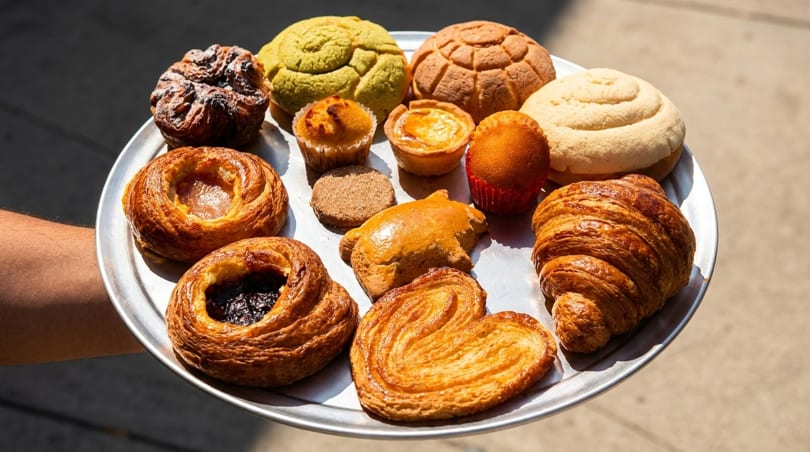 A person holds a silver tray filled with various pastries, including conchas, croissants, and palmiers.
