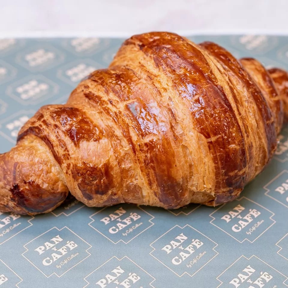 Golden-brown croissant with flaky layers, resting on blue Pan y Café branded paper.