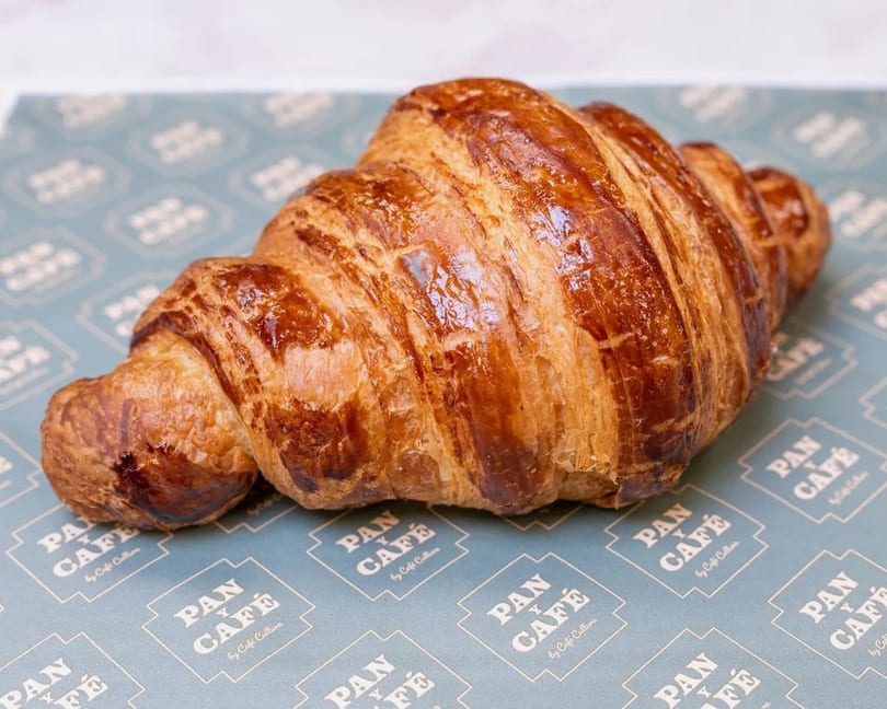 Golden-brown croissant with flaky layers, resting on blue Pan y Café branded paper.