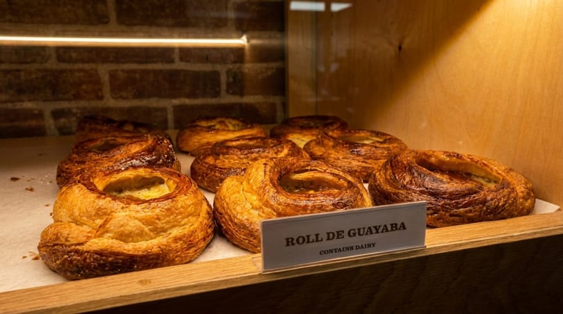 Golden-brown guava rolls with a flaky crust displayed in a wooden and brick bakery case.