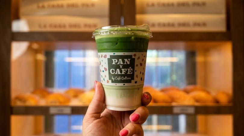 Hand holding a Pan y Café by Café Cultura matcha latte in front of a bakery display.