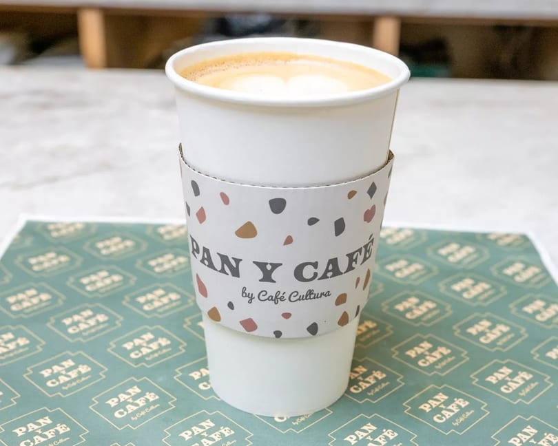 A Pan y Café by Café Cultura branded coffee cup with latte art sits on a branded placemat.