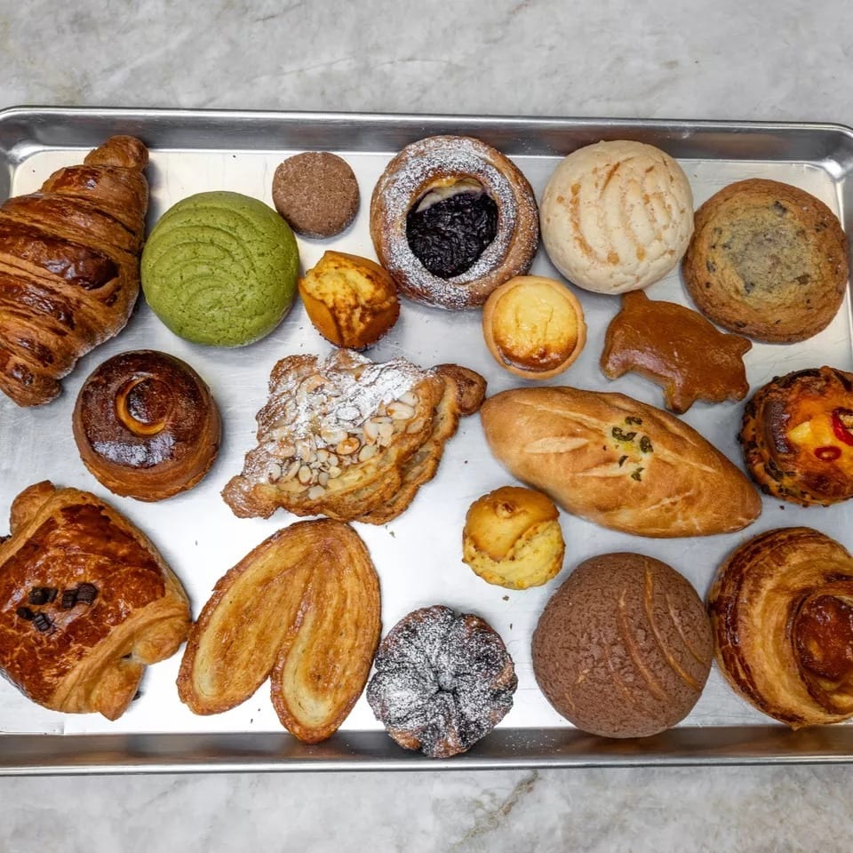 Assortment of pastries including croissants, conchas, danishes, and palmiers on a metal tray.