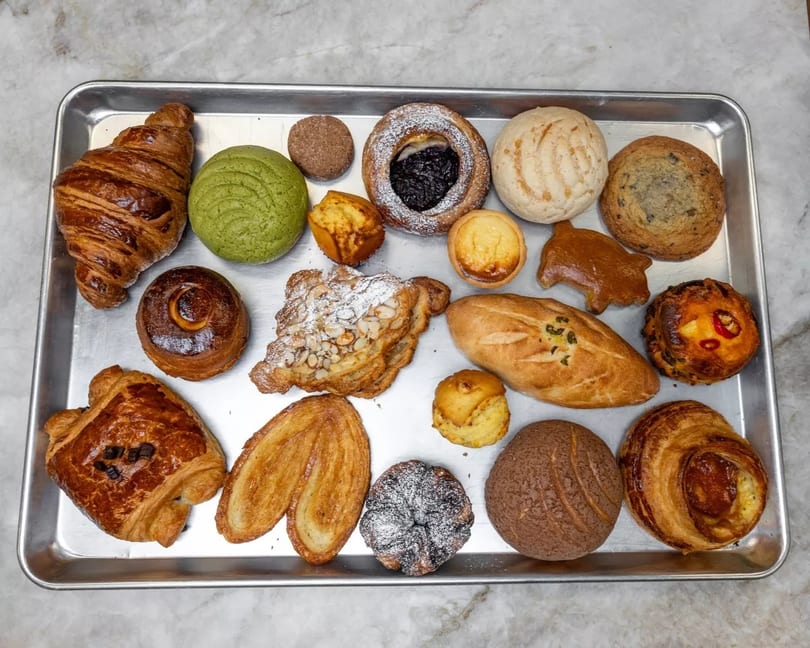 Assortment of pastries including croissants, conchas, danishes, and palmiers on a metal tray.