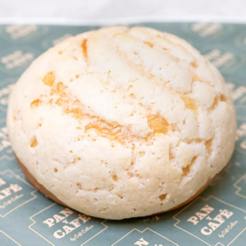 A close-up of a round, light-colored melon pan with a textured top on Pan y Café branded paper.