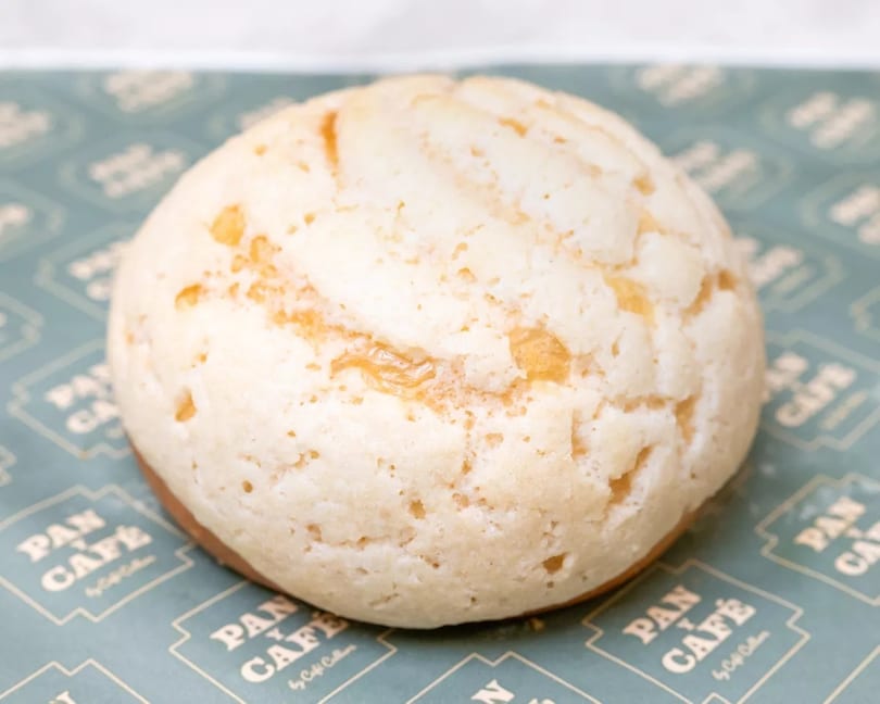 A close-up of a round, light-colored melon pan with a textured top on Pan y Café branded paper.