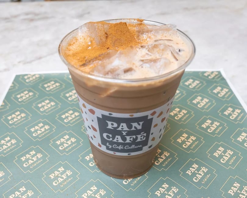 Iced coffee with cinnamon powder on top, in a Pan y Café branded cup and paper.