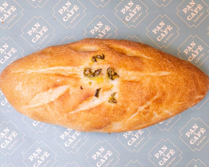 Golden-brown Pan y Café bread with green herbs on light blue branded paper.