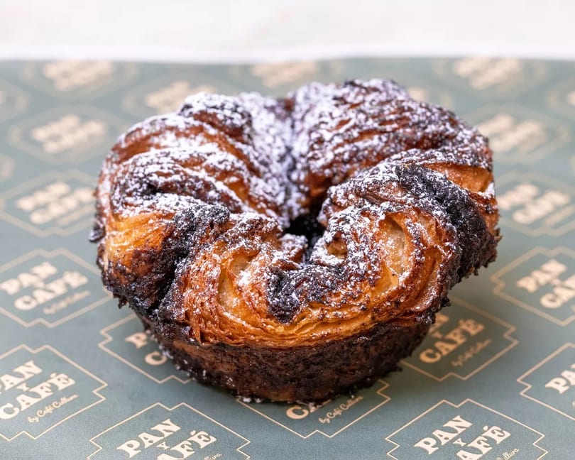 A chocolate croissant, dusted with powdered sugar, rests on a Pan y Café branded paper.