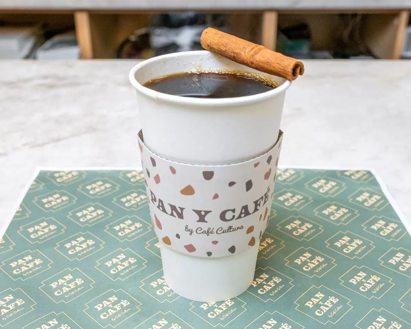 A Pan y Café coffee cup with a cinnamon stick on top, sitting on a branded placemat.
