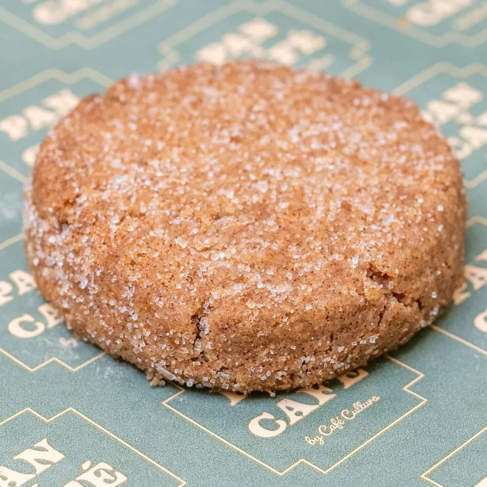 A round, brown cookie covered in sugar crystals rests on a green and gold patterned paper.