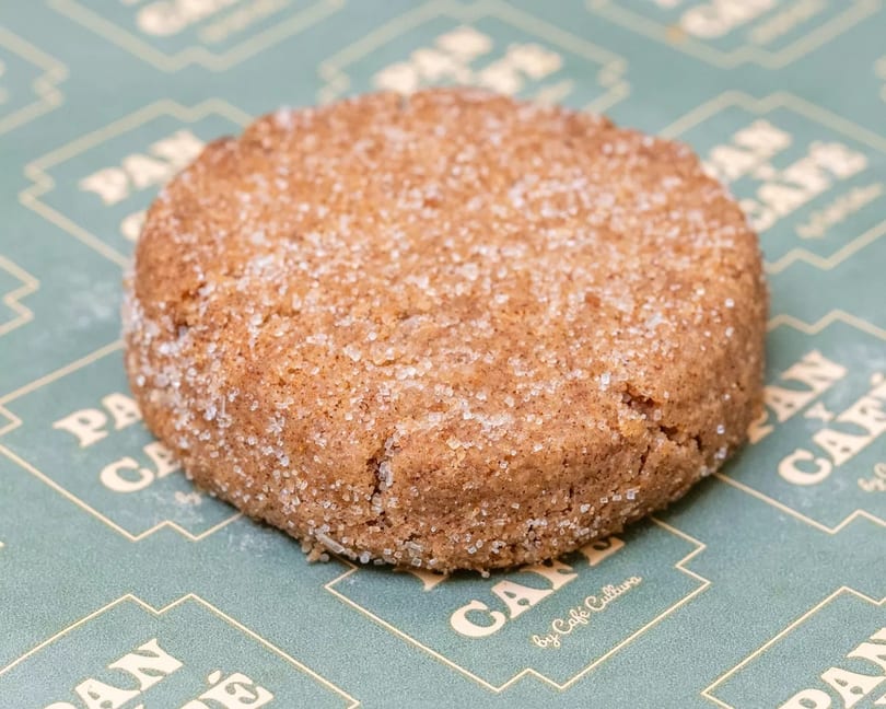 A round, brown cookie covered in sugar crystals rests on a green and gold patterned paper.