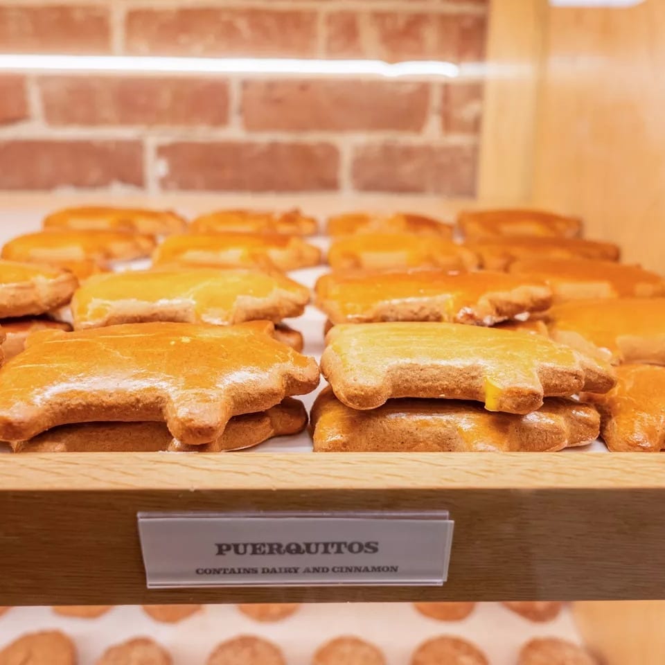 Rows of golden-brown puerquitos, pig-shaped cookies, displayed on a wooden shelf with a brick wall background.