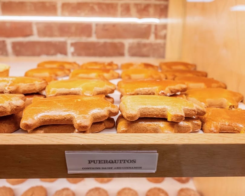 Rows of golden-brown puerquitos, pig-shaped cookies, displayed on a wooden shelf with a brick wall background.