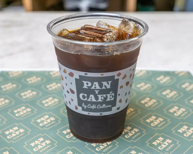 Iced coffee in a clear plastic cup with a "Pan y Café by Café Cultura" label, on a branded placemat.